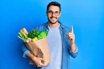 Young hispanic man holding paper bag with bread and groceries smiling with an idea or question pointing finger with happy face, number one