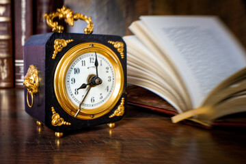 Old wooden alarm clock with gold applications, showing 8 o'clock on a wooden table with books in the background. Selective focus
