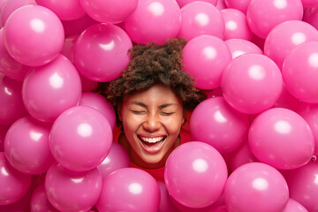 Fototapeta na wymiar Pretty overjoyed young woman has curly hair foolishes around smiles broadly with white teeth keeps eyes closed sticks out head through small pink balloons. Cheerful ethnic female enjoys party