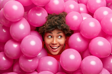 Beautiful happy curly dark skinned woman sticks head through heap of pink inflated balloons smiles...