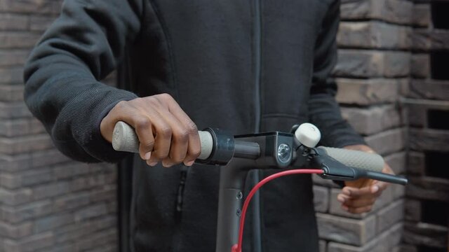 A young African-American in casual dark clothes with a scooter stands near a building with a dark brick facade and types on a smartphone. Close-up view of hands and steering wheel