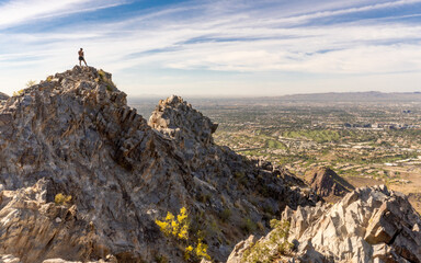 A lone hiker is seen atop Piestewa Peak in Phoenix, Arizona, on top of a rocky cliff overlooking the Valley of the Sun with a blue sky with high clouds.  - Powered by Adobe