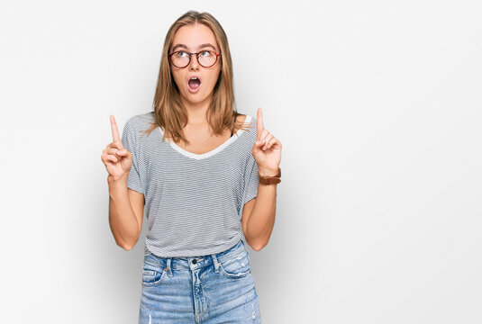 Beautiful young blonde woman wearing casual clothes and glasses amazed and surprised looking up and pointing with fingers and raised arms.