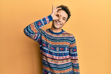 Young handsome man wearing casual winter sweater smiling happy doing ok sign with hand on eye looking through fingers