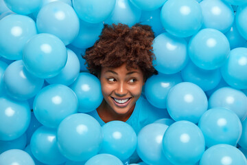 Fototapeta na wymiar Glad brunette young African American woman with curly hair sticks out head through inflated blue balloons smiles broadly has happy mood celebrates something. Holiday decor. Festive atmosphere.