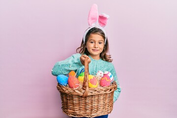 Little beautiful girl wearing cute easter bunny ears holding wicker basket with colored eggs...