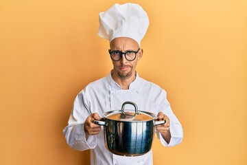 Bald man with beard wearing professional cook holding cooking pot skeptic and nervous, frowning upset because of problem. negative person.