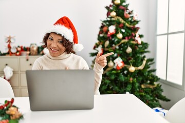 Middle age hispanic woman using laptop sitting by christmas tree smiling and looking at the camera pointing with two hands and fingers to the side.