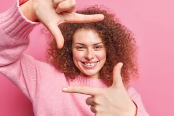 Beautiful cheerful young woman makes camera hand frame gesture smiles happily wears casual jumper...