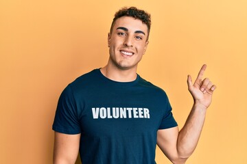 Hispanic young man wearing volunteer t shirt cheerful with a smile on face pointing with hand and finger up to the side with happy and natural expression