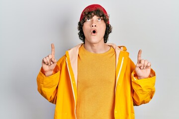 Handsome young man wearing yellow raincoat amazed and surprised looking up and pointing with fingers and raised arms.