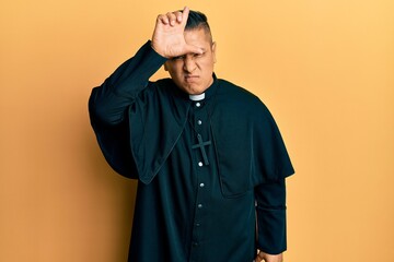 Young latin priest man standing over yellow background making fun of people with fingers on forehead doing loser gesture mocking and insulting.