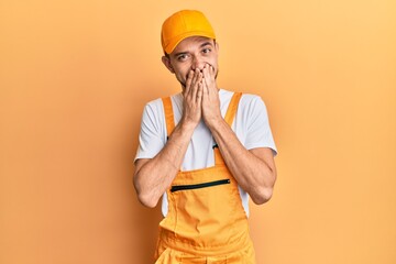Hispanic young man wearing handyman uniform laughing and embarrassed giggle covering mouth with...