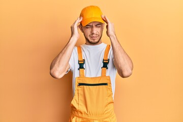 Hispanic young man wearing handyman uniform with hand on head, headache because stress. suffering migraine.