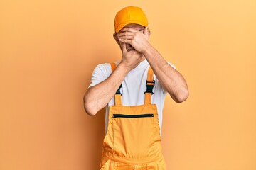 Hispanic young man wearing handyman uniform covering eyes and mouth with hands, surprised and...