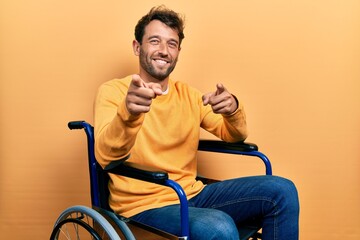 Handsome man with beard sitting on wheelchair pointing fingers to camera with happy and funny face. good energy and vibes.