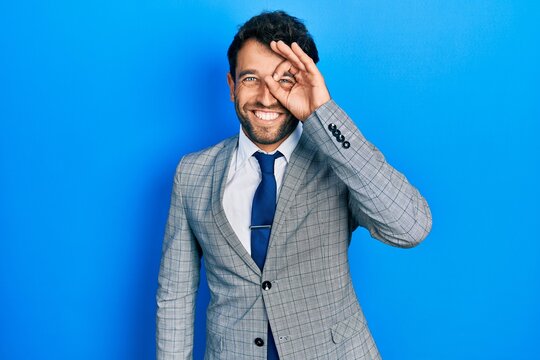 Handsome man with beard wearing business suit and tie doing ok gesture with hand smiling, eye looking through fingers with happy face.