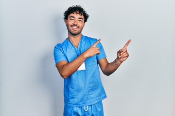 Young hispanic man wearing blue male nurse uniform smiling and looking at the camera pointing with two hands and fingers to the side.