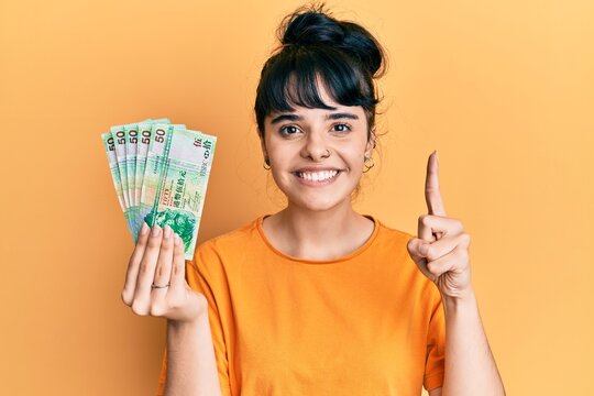 Young hispanic girl holding 50 hong kong dollars banknotes smiling with an idea or question pointing finger with happy face, number one