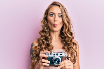 Young blonde girl holding vintage camera making fish face with mouth and squinting eyes, crazy and comical.