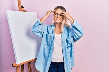 Beautiful young blonde woman standing by white painter easel stand with hand on head, headache because stress. suffering migraine.