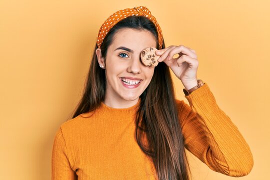 Young brunette teenager holding cookie over eye looking positive and happy standing and smiling with a confident smile showing teeth