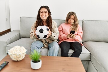Girl smiling happy watching soccer match and her girlfriend boring using smartphone at home.