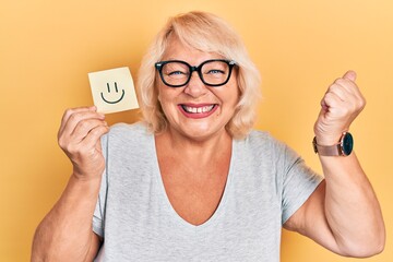 Middle age blonde woman holding smile reminder screaming proud, celebrating victory and success very excited with raised arm
