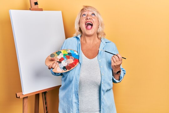 Middle age blonde woman standing drawing with palette by painter easel stand angry and mad screaming frustrated and furious, shouting with anger looking up.