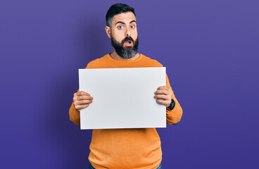 Hispanic man with beard holding blank empty banner in shock face, looking skeptical and sarcastic, surprised with open mouth