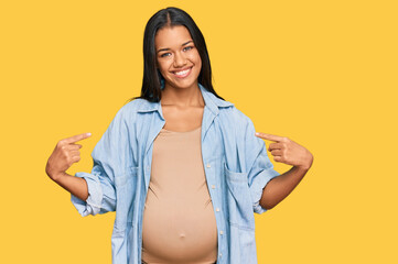 Beautiful hispanic woman expecting a baby showing pregnant belly looking confident with smile on...