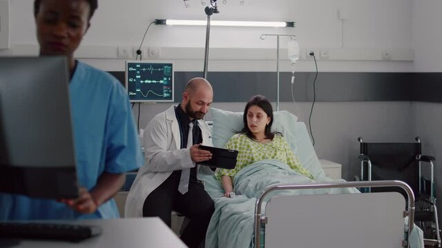 Practitioner doctor explaining disease diagnosis to sick woman using tablet computer. Patient resting in bed recovering after surgery in hospital ward while black nurse typing medical expertise