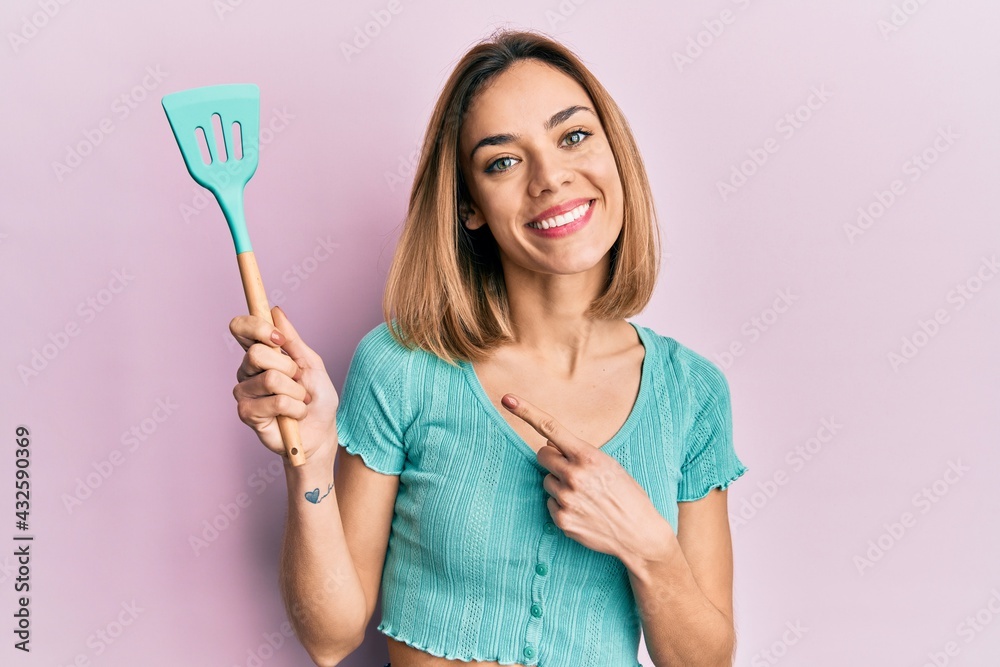 Wall mural young caucasian blonde woman holding silicone spatula smiling happy pointing with hand and finger