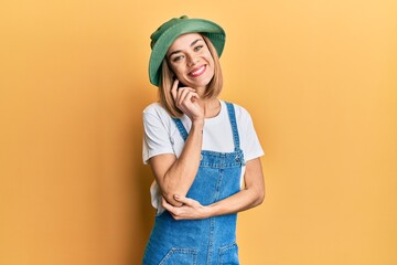 Young caucasian blonde woman wearing denim jumpsuit and hat with 90s style looking confident at the...
