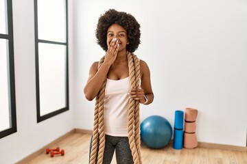 Young african american woman with afro hair at the gym training with battle ropes laughing and...