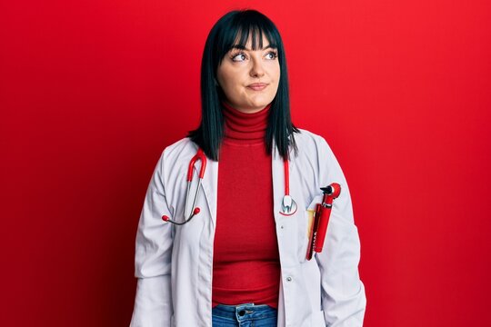 Young hispanic woman wearing doctor uniform and stethoscope smiling looking to the side and staring away thinking.