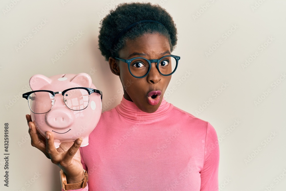 Sticker Young african american girl holding piggy bank with glasses scared and amazed with open mouth for surprise, disbelief face