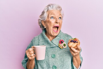 Senior grey-haired woman drinking a cup of coffee and eating bun angry and mad screaming frustrated...