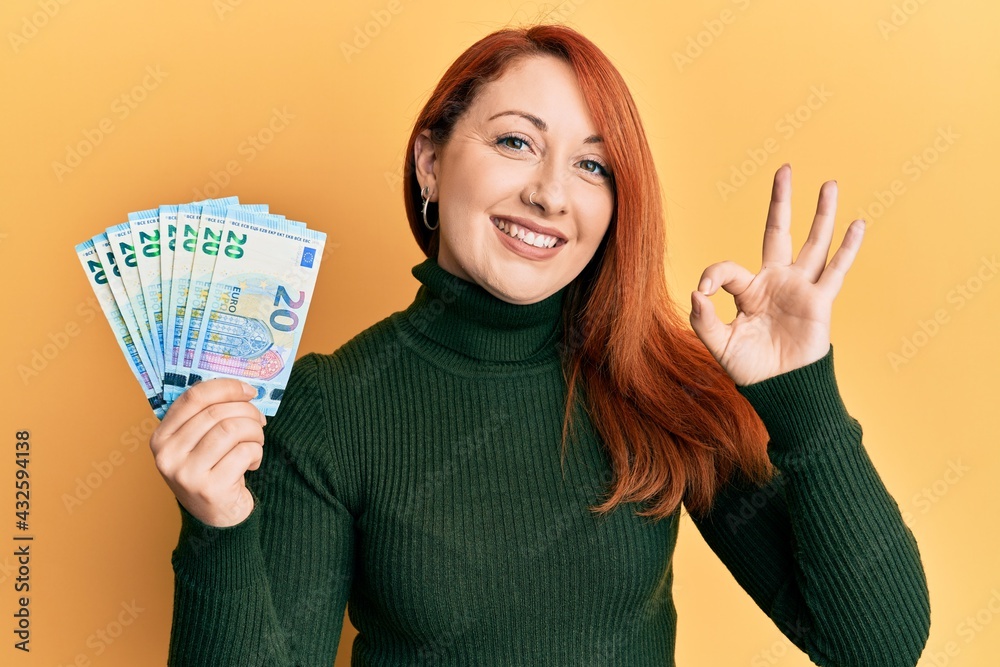 Canvas Prints beautiful redhead woman holding bunch of 20 euro banknotes doing ok sign with fingers, smiling frien