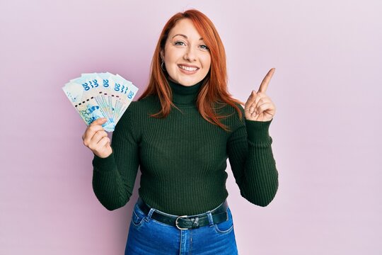 Beautiful redhead woman holding 20 hong kong dollars banknotes smiling happy pointing with hand and finger to the side