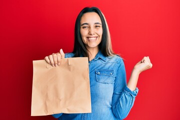 Young hispanic woman holding take away paper bag screaming proud, celebrating victory and success very excited with raised arm