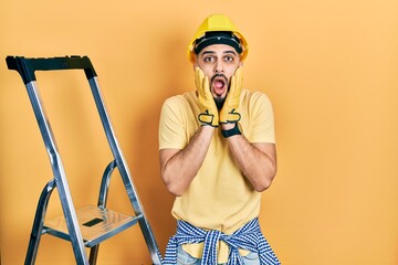 Handsome man with beard by construction stairs wearing hardhat afraid and shocked, surprise and amazed expression with hands on face