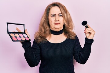 Middle age caucasian woman holding makeup brush and blush skeptic and nervous, frowning upset...