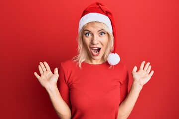 Young blonde girl wearing christmas hat celebrating victory with happy smile and winner expression with raised hands