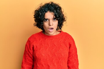 Young hispanic woman with curly hair wearing casual winter sweater in shock face, looking skeptical and sarcastic, surprised with open mouth
