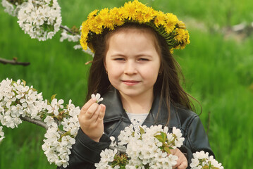 Portrait of a little girl in a wreath of dandelions near flowering trees