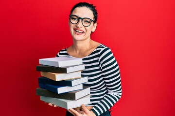 Young caucasian woman wearing glasses and holding books looking positive and happy standing and smiling with a confident smile showing teeth