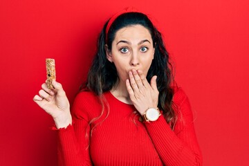 Young hispanic woman eating protein bar as healthy energy snack covering mouth with hand, shocked...