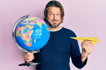 Fototapeta premium Middle age handsome man holding paper plane and world ball puffing cheeks with funny face. mouth inflated with air, catching air.