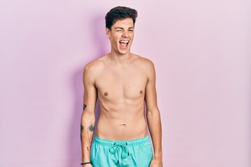 Fototapeta premium Young hispanic man wearing swimwear shirtless winking looking at the camera with sexy expression, cheerful and happy face.